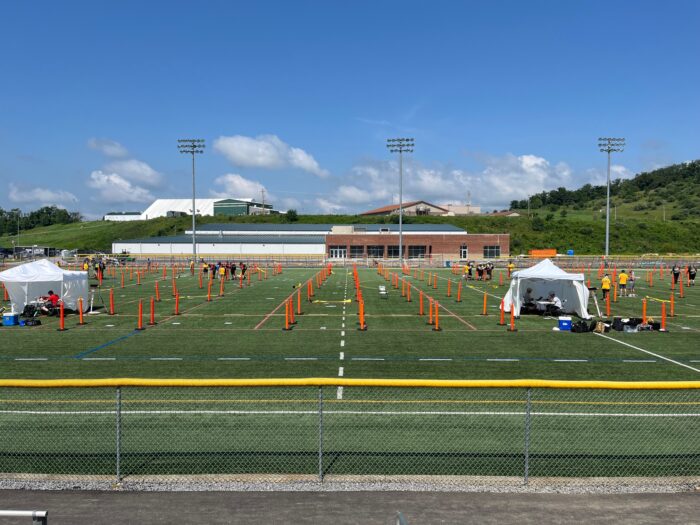 A soccer field covered in green turf under a clear sky on a summer day is arranged with orange barriers in between two white tents.