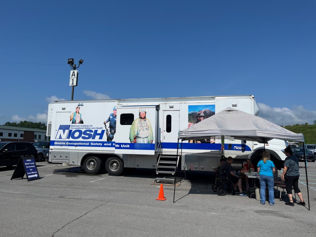 A large white truck with blue lettering sits in a parking lot with a tent sheltering multiple people from the sun on a hot day.