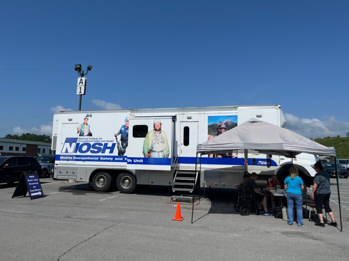 A large white truck with blue lettering sits in a parking lot with a tent sheltering multiple people from the sun on a hot day.