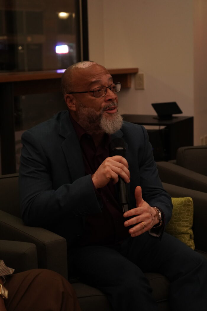 A Black man sits in a chair and speaks into a microphone. He is bald with a gray beard and wears glasses. He is formally dressed in a black suit.