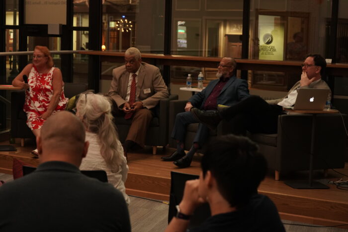 Four adults sit in chairs on a stage looking out at the audience. One white woman, one white man, and two Black men. They are all formally dressed. Two wear glasses.