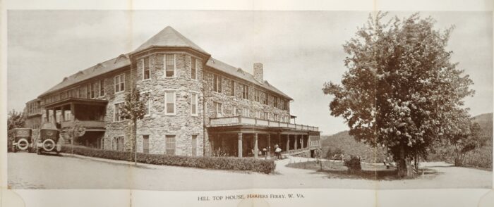 A historic, sepia-tinted photo shows a stone building with a small spire and expansive porch on a hillside. A tree and dirt road sit out front, and some people are visible on the porch in the distance.