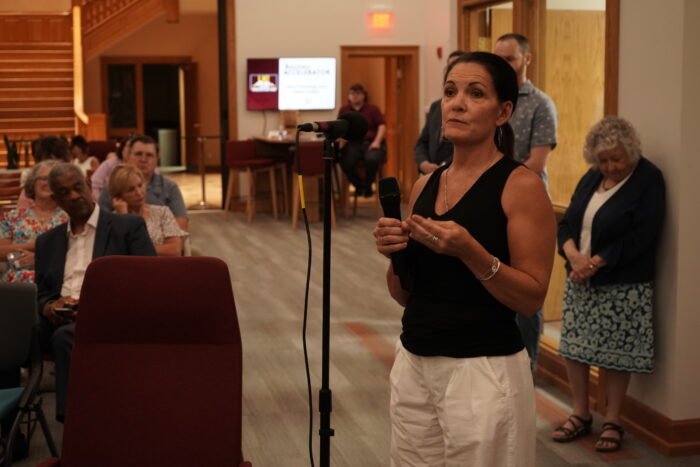 A white woman with dark hair speaks into a microphone. Her hair is pulled back in a low ponytail. She wears a black, sleeveless shirt and white pants.