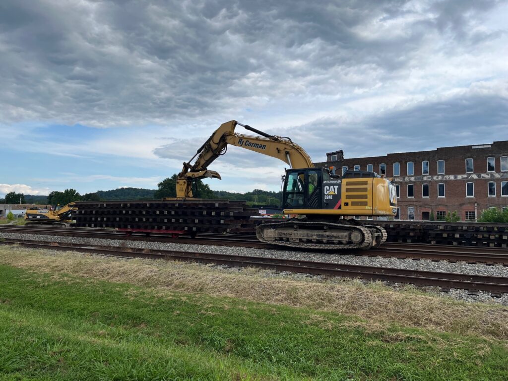 A yellow piece of construction machinery pushes a stack of prefabricated track panels down a railroad track under an overcast sky.