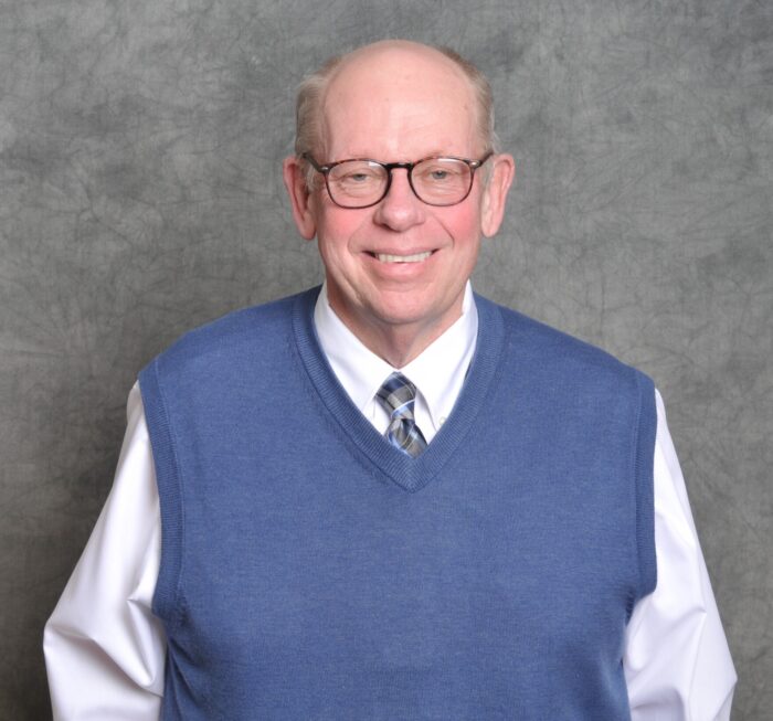 An older white man wearing glasses smiles for the camera. He wears a blue sweater over a white button up shirt and blue tie.