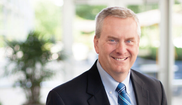 An older white man with gray hair smiles for the camera for a professional headshot. He wears a black blazer with a blue tie.