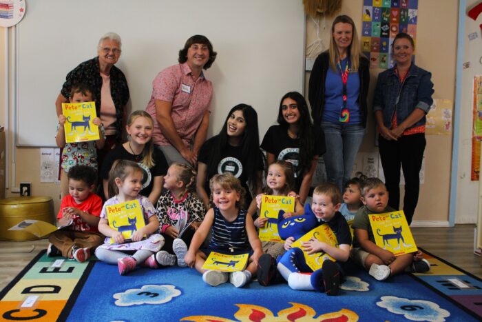 A group of children hold paperback books with yellow covers, seated on a colorful blue area rug. Behind them sit young women wearing black shirts, flanked by adults.