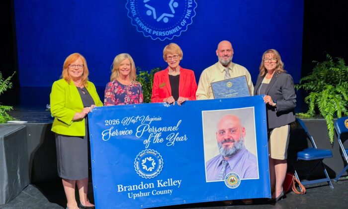 Four women and one man stand together on a stage in front of a blue screen displaying a seal that reads "West Virginia Teacher of the Year". In front of the women is a blue banner with script that reads "2026 West Virginia School Service Personell of the Year Brandon Kelley Upshur County" with the same seal in the middle and an image of the man.