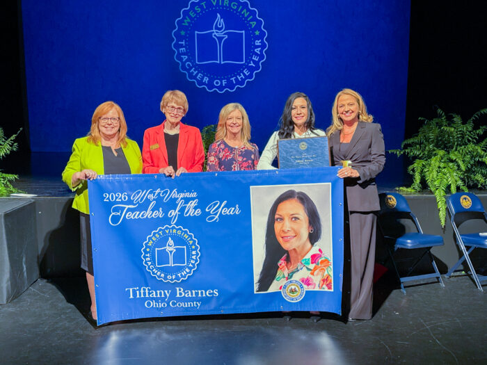 Five women stand together on a stage in front of a blue screen displaying a seal that reads "West Virginia Teacher of the Year". In front of the women is a blue banner with script that reads "2026 West Virginia Teacher of the Year Tiffany Barnes Ohio County" with the same seal in the middle and an image of the woman second from the left.