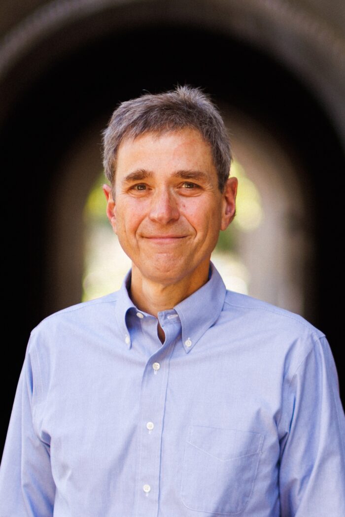 A professional photograph of a white man with gray hair. He wears a light blue button-up shirt. He looks directly into the camera.