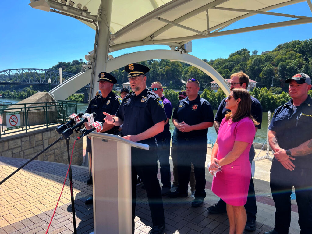 A man in a blue police uniform is flanked by other police officials in uniform and a woman in a hot pink dress wearing sunglasses.