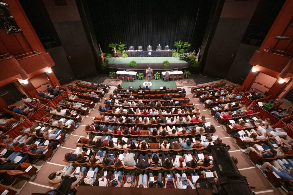 A balcony view in a theater with five men and women seated on stage