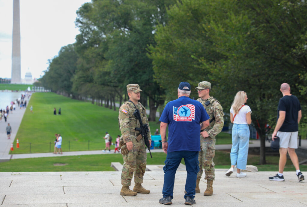 two men in uniform speak to a man in a t-shirt on the national mall with the Washington Monument in the background.