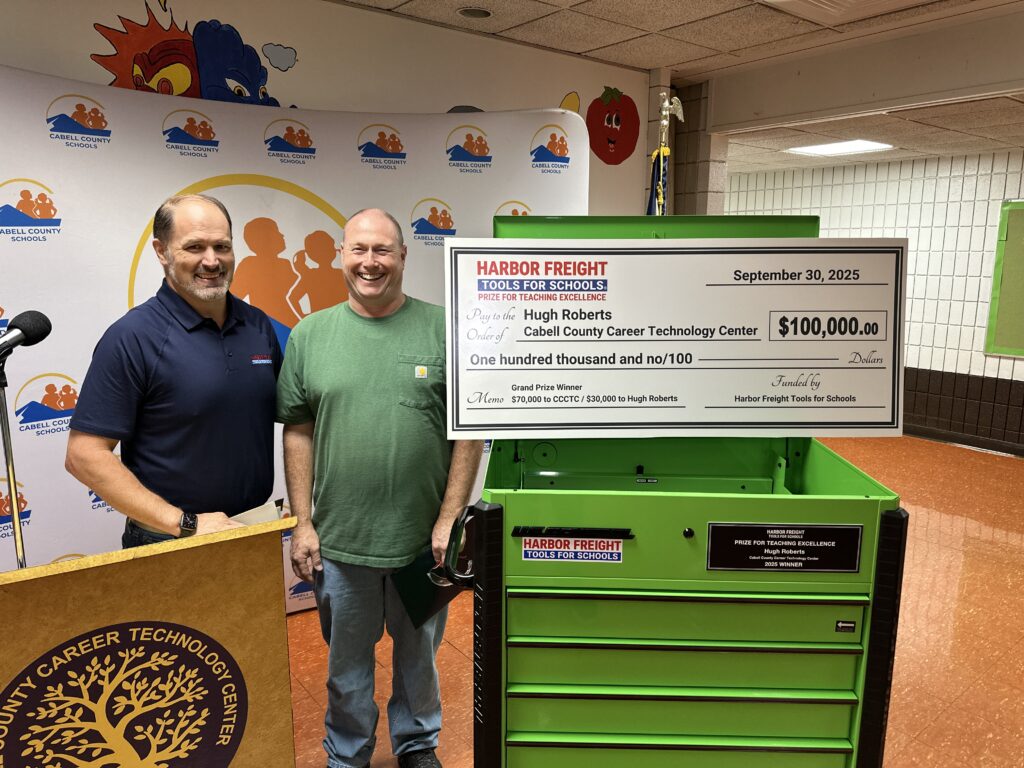 A man in a blue polo shirt stands next to a smiling bald man in a green t-shirt and jeans standing next to a novelty check for $100,000 propped up on a lime green tool chest in a classroom.