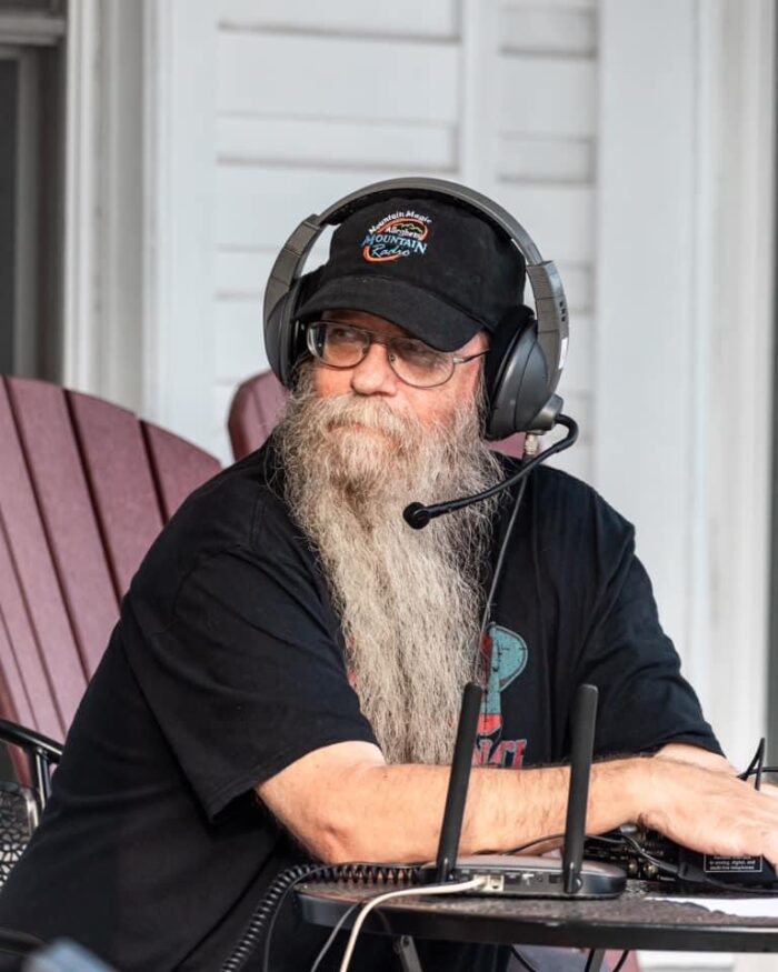 A white man with a white beard sitting at a desk wearing a hat and over-the-ear headphones. He is looking away from the camera.
