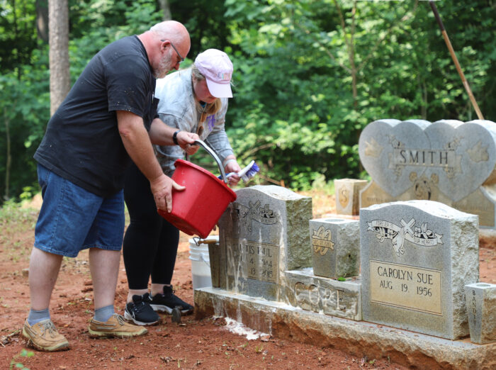 Two people pouring a bucket of soap and water over a tombstone in a cemetery. 