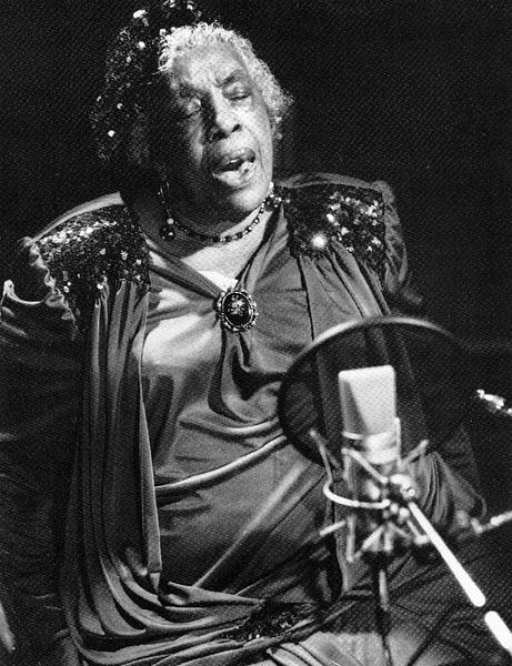 A black and white photo of a Black woman singing into a microphone.