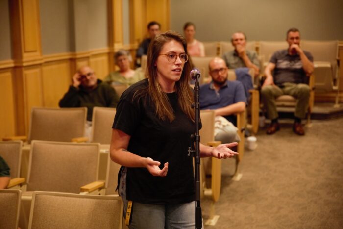 A millennial age woman with long brown hair and wearing glasses speaks into a standing microphone from the audience.