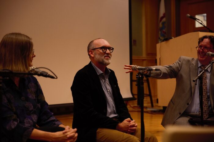 A balding man sits on the lip of a stage and looks up as if thinking. He wears glasses and speaks to the audience from a microphone.