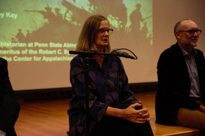 A woman sits on the lip of a stage with her hands clasped around one knee. She speaks in a microphone.