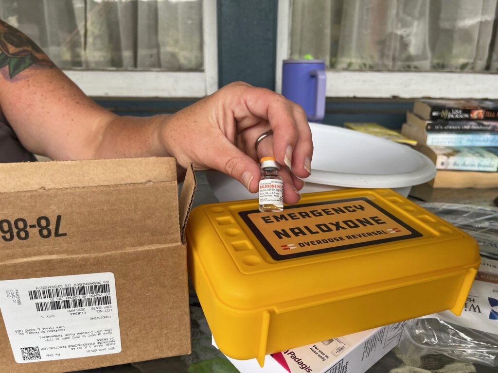 A woman with a tattooed arm holds a small glass vial above a bright yellow plastic box on a table filled with overdose treatments.