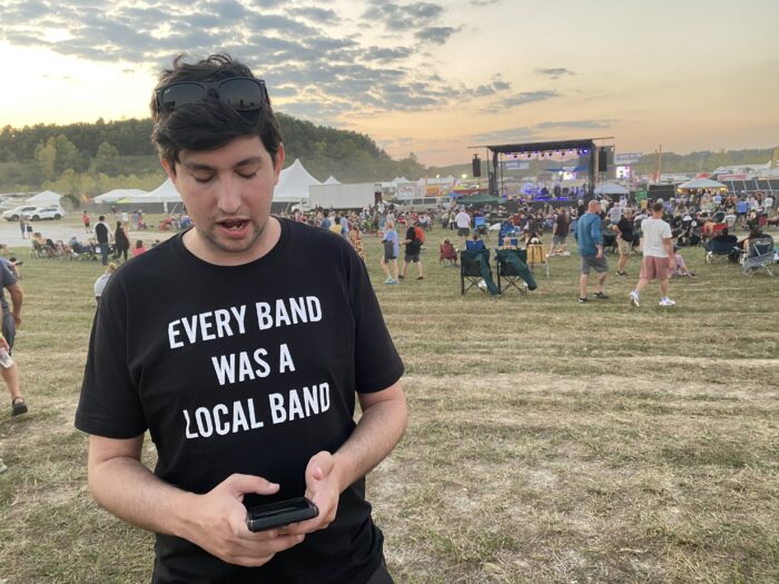 Man in a field in front of a stage, wearing a t-shirt that says