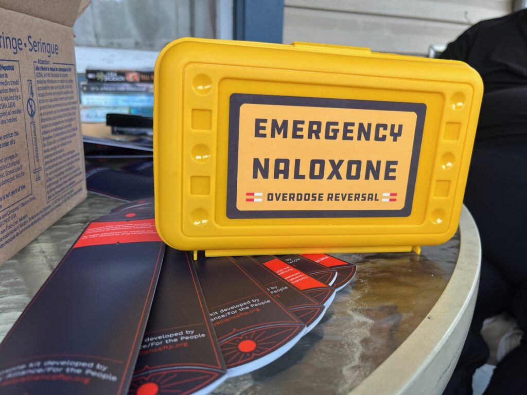 A yellow box with a label that says 'Emergency Naloxone' sits on top of red and blue sleeves to hold needles and naloxone on a table on a porch.