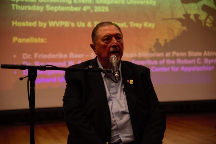 An older man sits on the lip of a stage and speaks into a microphone.
