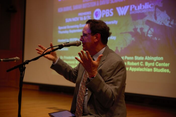 A man speaks to an audience while sitting on the lip of a stage.