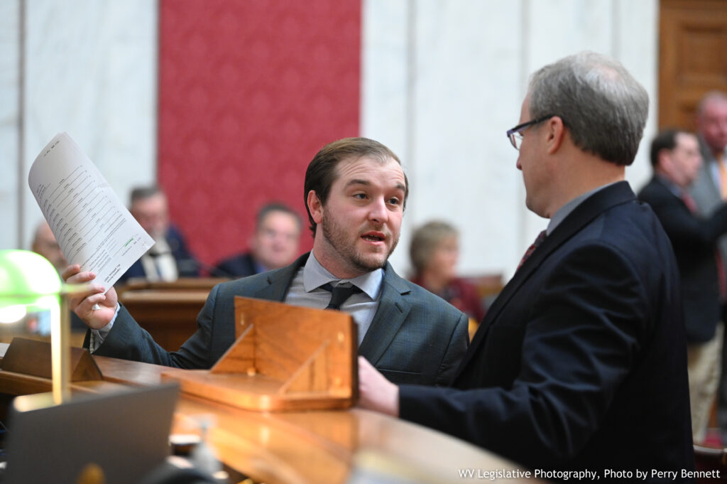 A man in a grey suit holds up a sheaf of papers while standing at a wooden rail. To his right stands a man in a dark suit facing the first man.