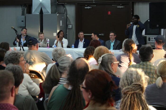 A panel of men and one woman wearing blazers over button up shirts sit behind a long table covered with a white tablecloth. The man on the far right of the panel is standing and holding a microphone up to his mouth. In front of them stretching to the forefront is a seated crowd.