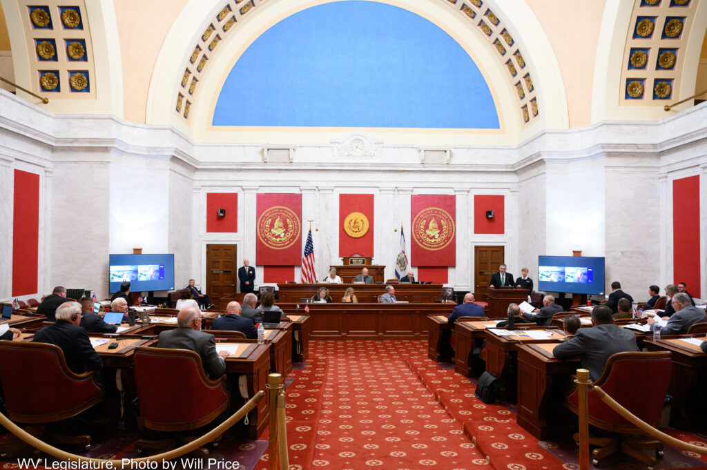 A valuted chamber with ornate red carpet has an array of wooden desks facing a dark wood dais in front of a wall of white marble accented by red and state seals.