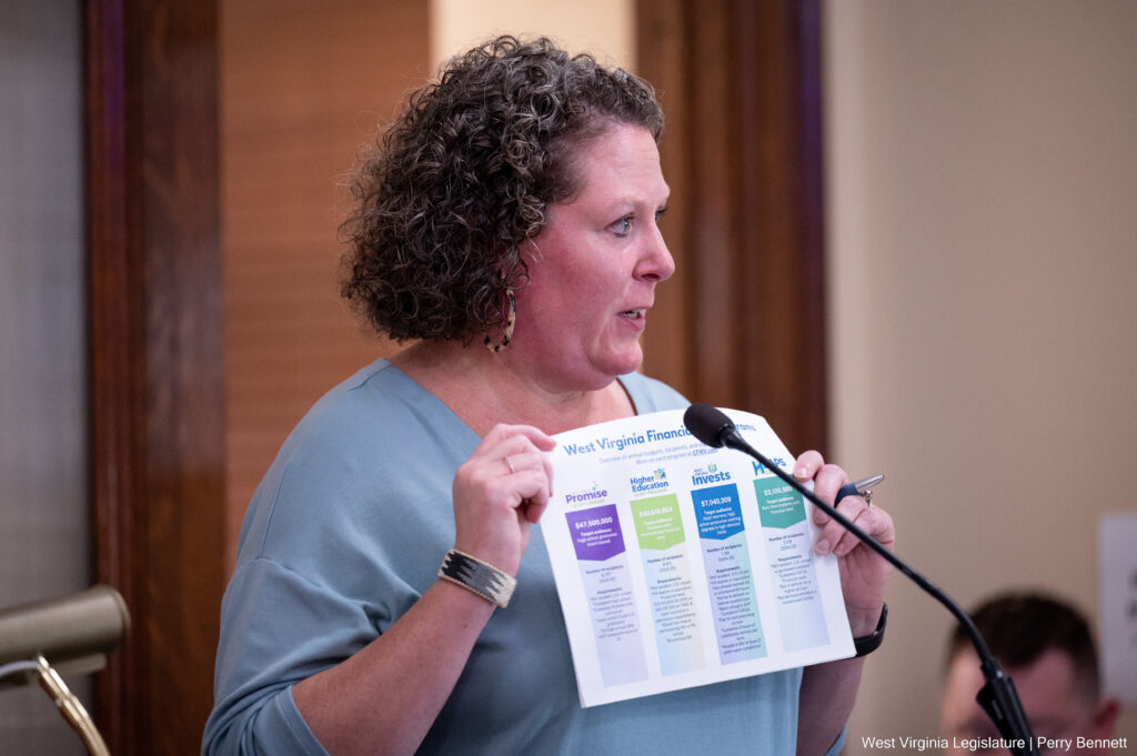A woman with curly hair and a light blue blouse holds up a chart behind a microphone.