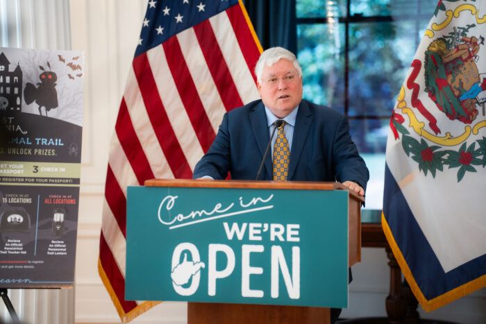A man in a dark blue suit over a light blue shirt and gold tie stands behind a lectern with a sign hung on it that reads "Come in we're open." The man is standing in front of a U.S. flag and a West Virginia flag. To his right is the portion of a poster with a cartoon mothman on it.