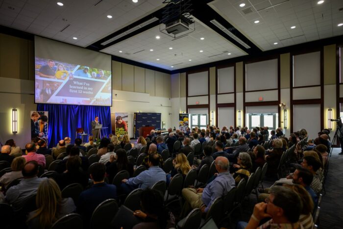 A large gathering of seated people faces towards a large projector screen. A man stands at a lectern below the screen and in front of a blue backdrop.