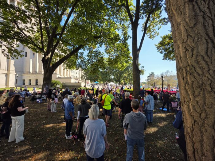 A large crowd stands between trees to the side of the riverside entrance of the West Virginia Capitol. Beyond the trees can be seen the white stone of the columned building.