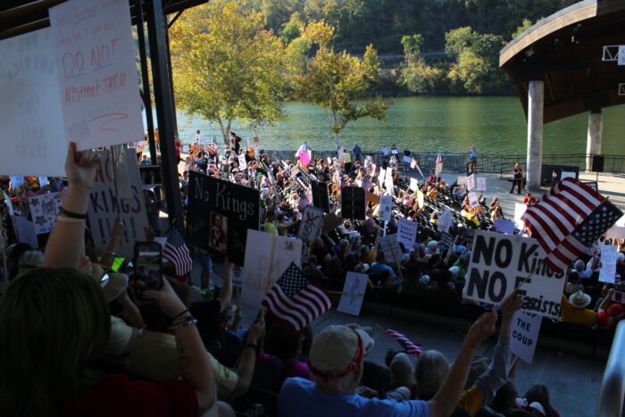 A large crowd seated in in an amphitheater by a broad river hold up protest signs.