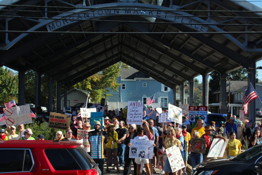 A large crowd stands in a covered outdoor patio holding up protest signs. Cars cross the frame in front of the crowd.