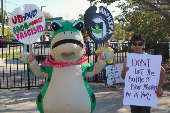 A person dressed in an inflatable frog costume holds up circular protests signs. The left sign reads "Unpaid Frog Against Fascism" and the one on the right reads "Amphifa: Amphibians Against Fascism" To the frog's right a man holds a white poster board sign that reads "Don't let the Battle of Blair Mountain be in vain!"