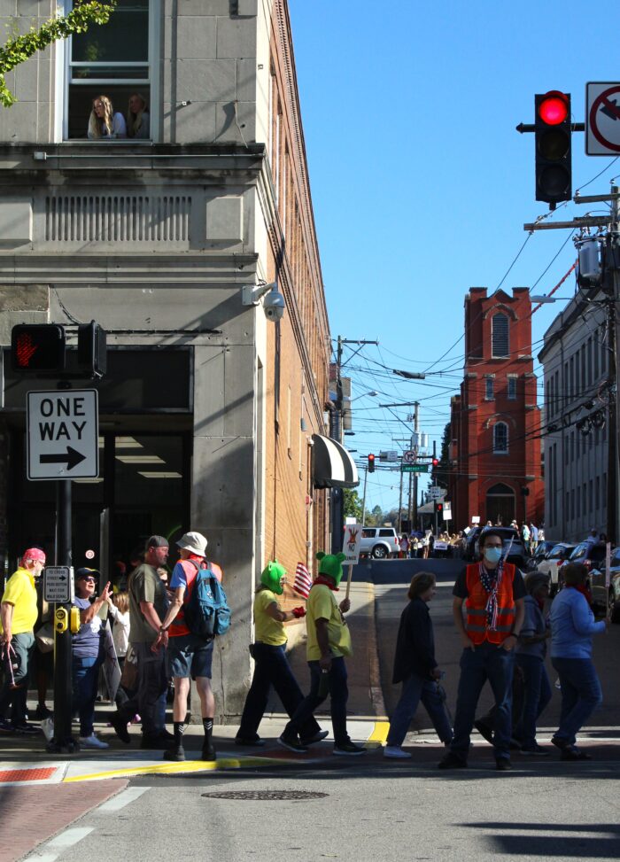 A group of people cross the street behind ushers wearing orange vests. Above them a group of young women look down on the marchers. Down an alley, more demonstrators from the same group can be seen on a parallel street.