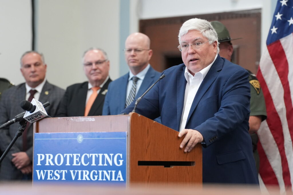 A man wearing a dark blue suit jacket over a white button down collared shirt with no tie grasps the edges of a wooden lectern with a sign that reads "Protecting West Virginia" on its front. Behind him stand several suited men.