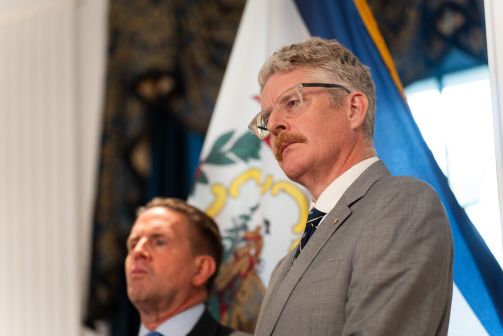 Two men wearing suits and ties stand in front of a West Virginia flag. The man on the left wears a dark suit, the man on the right wears glasses and a grey suit.