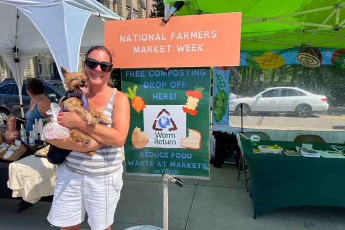 A woman holding a dog in at a farmers market in front of a sign that reads, "National Farmers Market Week. Free composting. Drop off here."