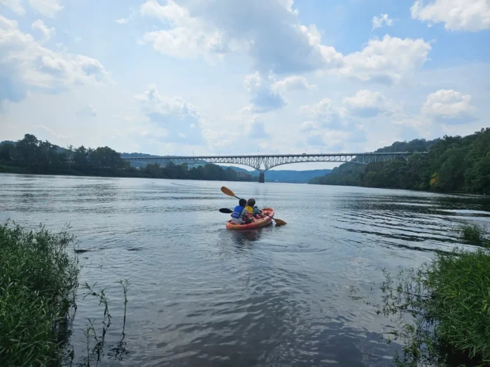 Two people sitting in a kayak on a river. There is a bridge in front of them.