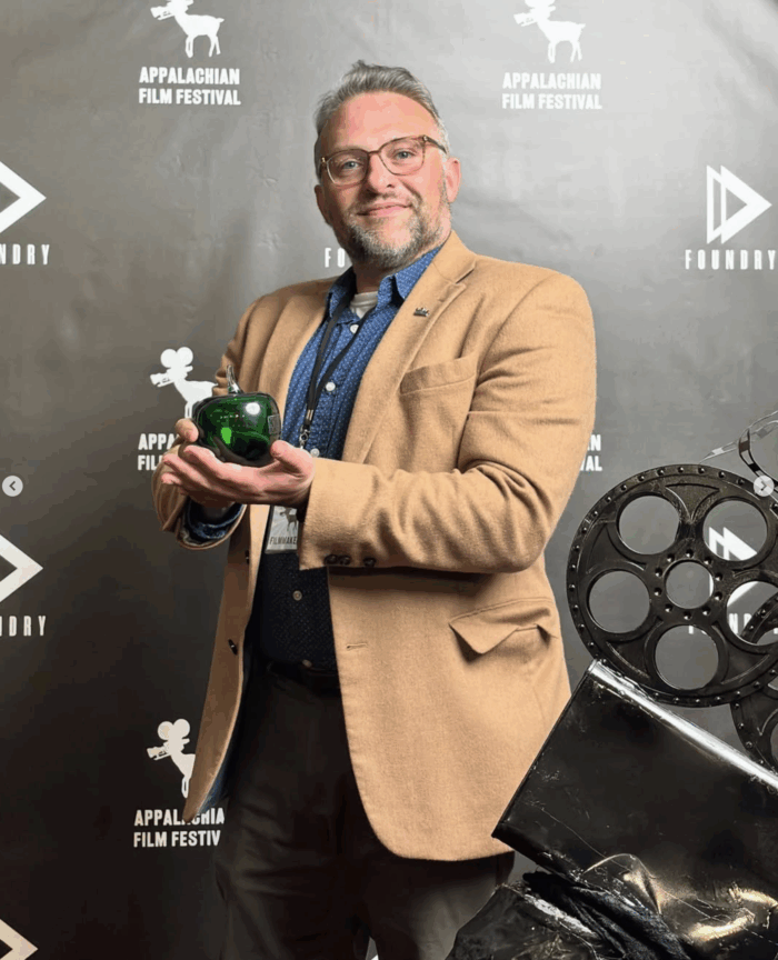 A white man holding a green glass apple. He is standing in front of a black backdrop and there is a film reel in the bottom right corner of the photo. 