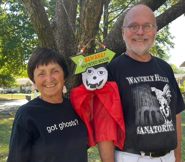 Two white people, a woman and a man, standing with a scarecrow-like figure between them.