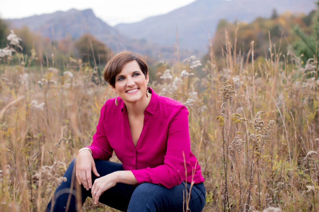 A woman sits in a field smiling and look up off camera. She has short hair and wears a pink shirt and jeans.