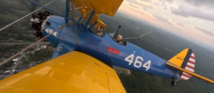 A yellow and blue plane in the air with two men in the separate open-air cockpits.