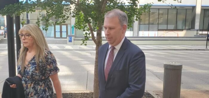 A gray haired man wearing a suit and tie walks outside a federal courthouse accompanied by a woman with blonde hair.
