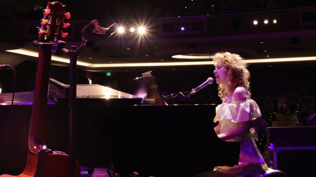 A woman with blonde hair and a white dress sits at the piano and plays while she sings into a mic.